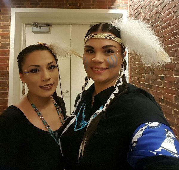 Heads and torsos of two women smiling. They wear black tops, necklaces, stage face makeup designs, and a feather in their hair. One has a headband.