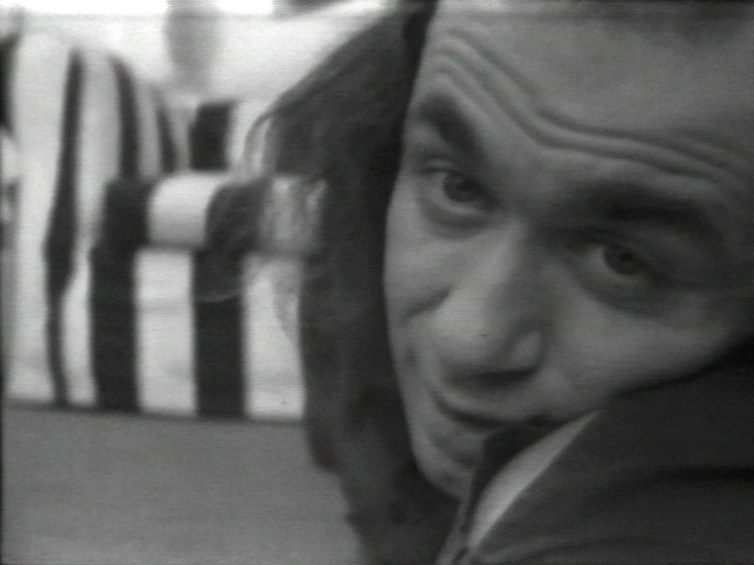 Black and white still of Vito Acconci lying on the ground, staring intently at the camera. A couch with vertical stripes is visible behind him.