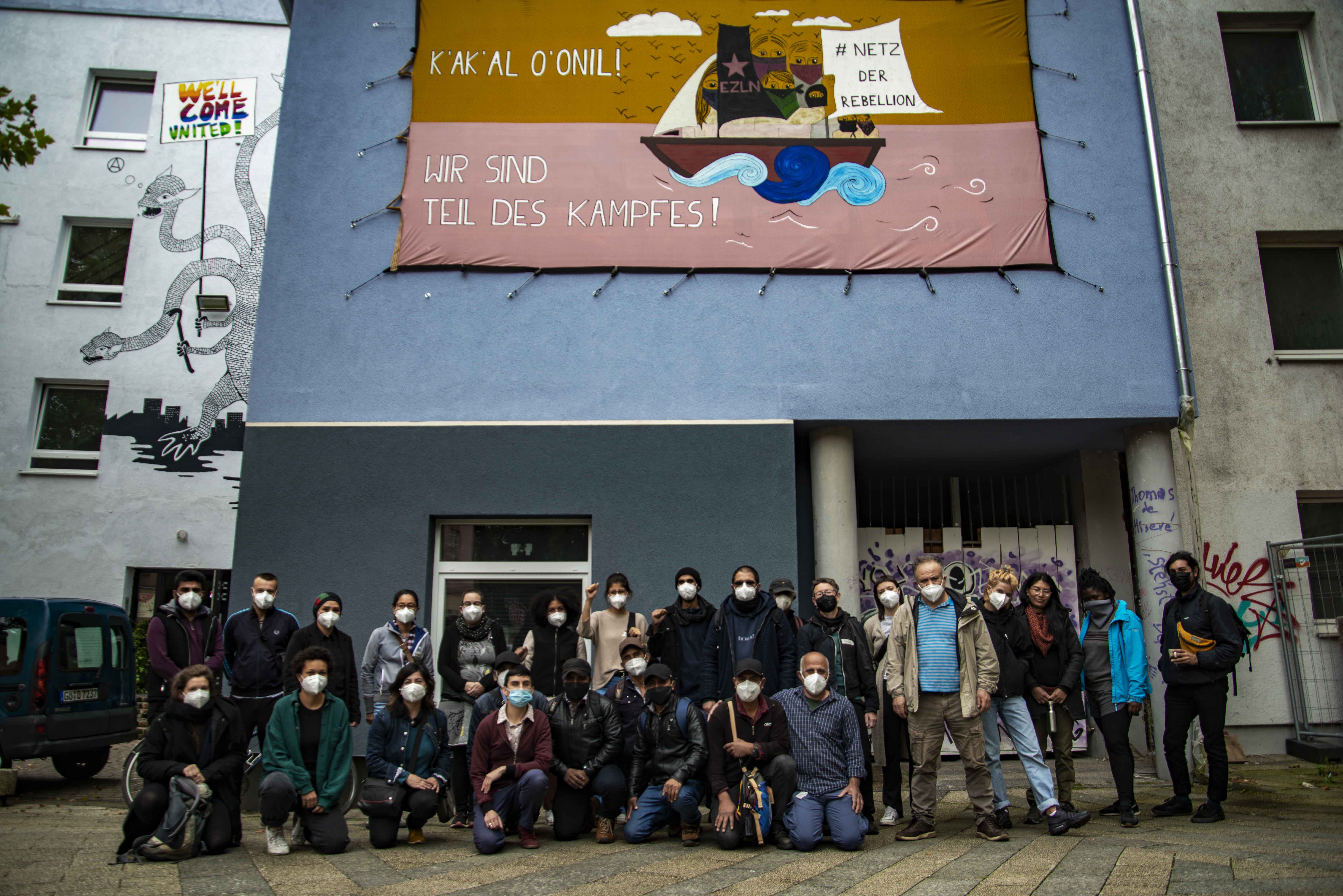 The blue wall of a building is covered by a large banner with the words “K'AK'AL O'ONIL! Wir sind Teil des Kampfes!” (We are part of the struggle). On the banner, there is the image of a boat with members of the EZLN (Zapatista Army of National Liberation) wearing masks and holding a sign reading “Network of Rebellion.” In front of the building there is one row of people from a collective gathering standing and one row of people kneeling. Most people are wearing medical masks.