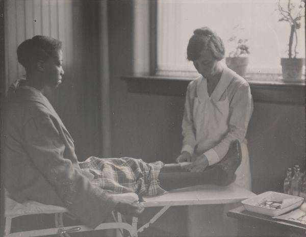 A young African American woman sits on a gurney table while a White nurse applies a bandage to her shin.