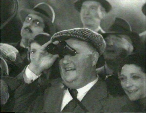 A crowd of happy people stares intently at something ahead of them. A man in the center holds binoculars to his face.