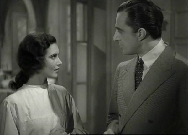 A tall man dressed in a suit and a young woman in a white dress, both in profile, gaze into each other’s eyes as they converse.