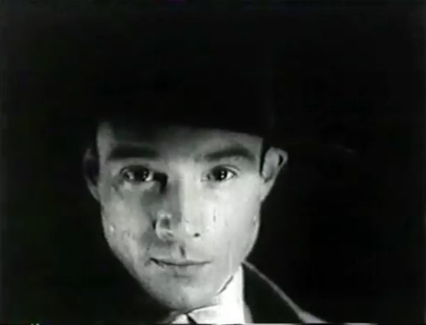 The face of a young man dominates the scene as he stares directly and smirks deviously at the camera.