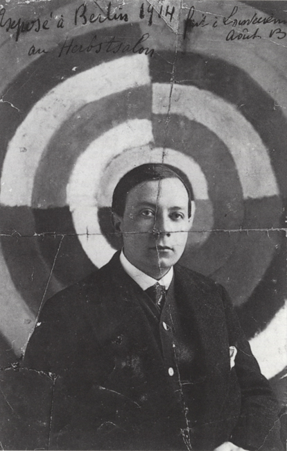 The exhibit features a photograph capturing Robert Delaunay standing before his painting, “The First Disk.” The artwork takes on the appearance of a target, and Robert is intentionally positioned right in front of its center. He is dressed in formal attire, wearing a coat.