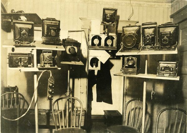Empty room with four wooden chairs situated in front of series of camera stands. Each stand is laden with four or more accordion-style cameras.