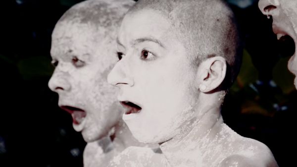 Two figures covered in white makeup with open mouths; the mouth of a third figure can be seen on the periphery.