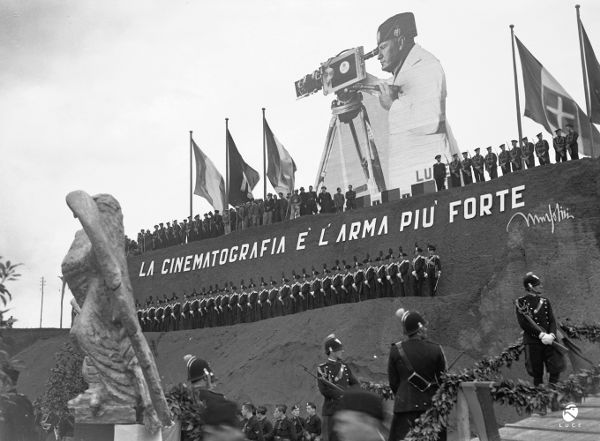 A gigantic cutout of Benito Mussolini peers into a movie camera, looming over the uniformed crowds lined up at the headquarters of the LUCE Institute.
