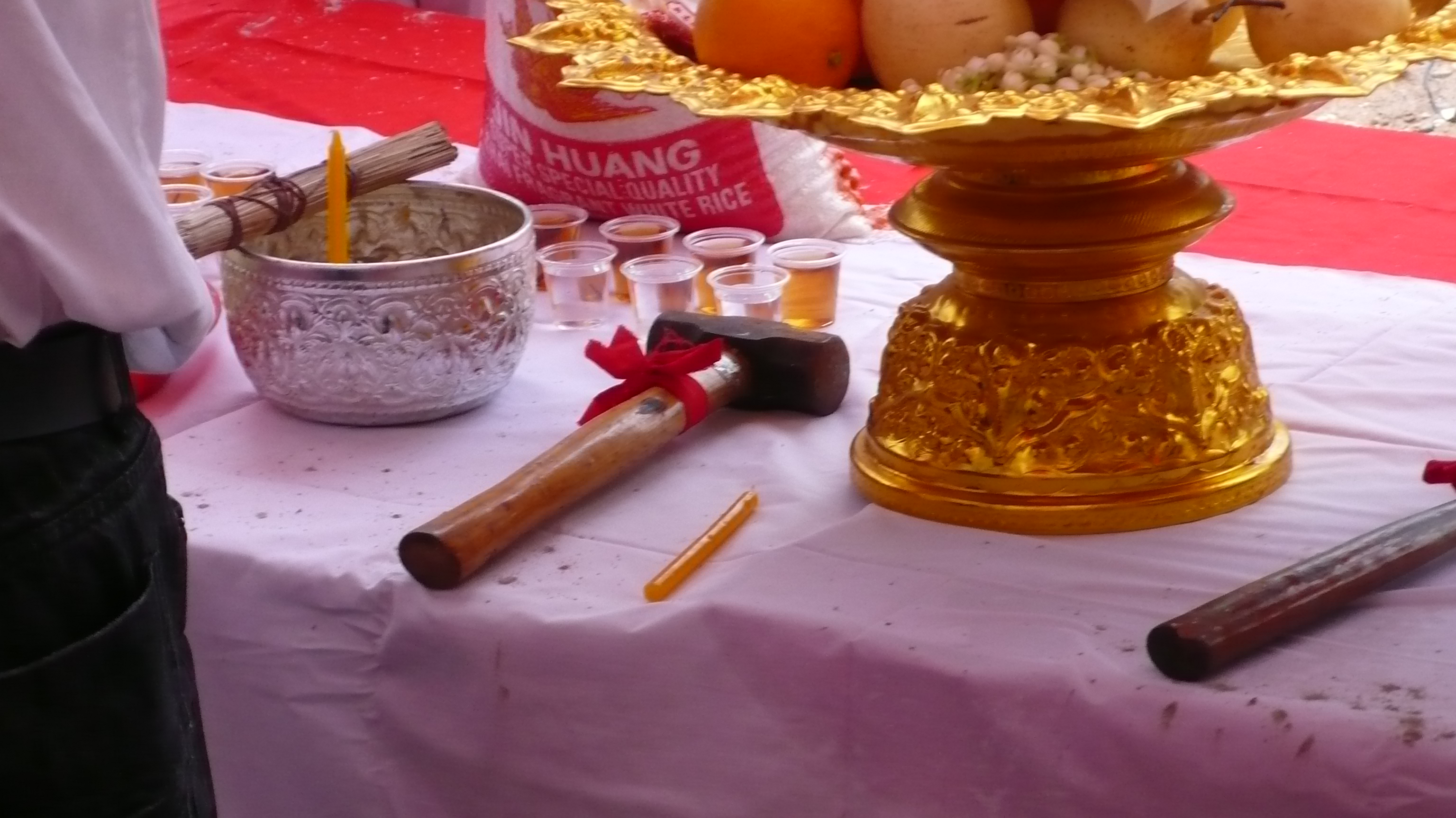 A table is covered in white cloth. On its surface stand an ornamental bowl and platter. There is a hammer, with its handle tied in red ribbon.
