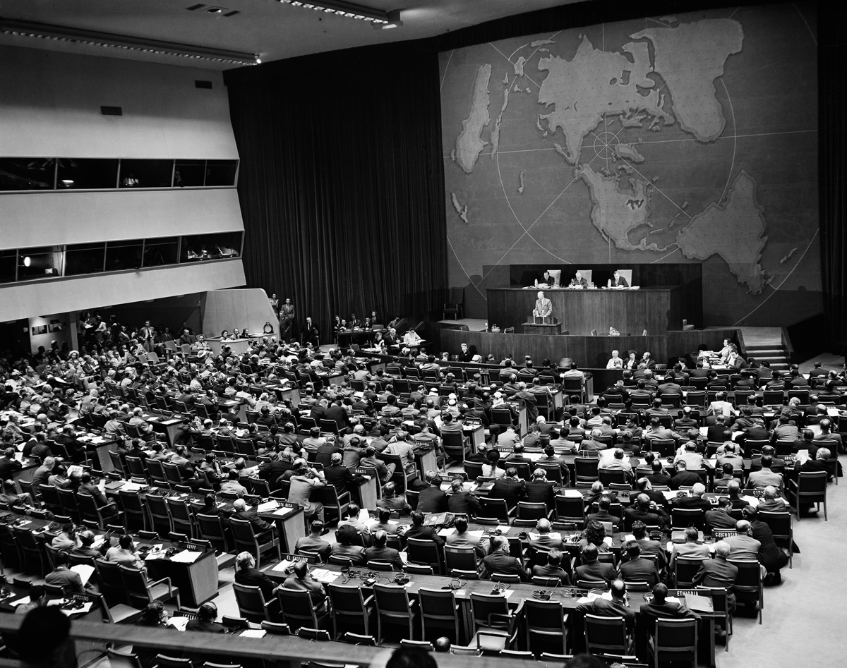 Photograph of interior of the plenary hall with seated delegates, podium, the presidential rostrum and media booth galleries on one side.