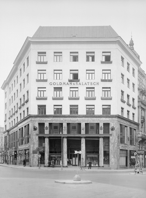 The exhibit displays the Goldman and Salatsch store from a distance, showcasing a substantial five-story building. The structure appears large, and there are people roaming about in the vicinity.