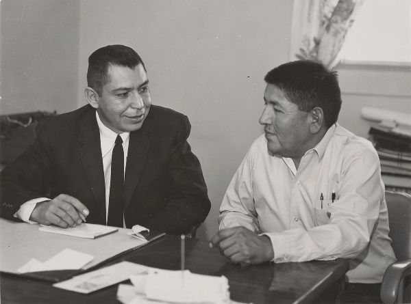 Two men sit behind an office desk, smiling at each other; one takes notes.
