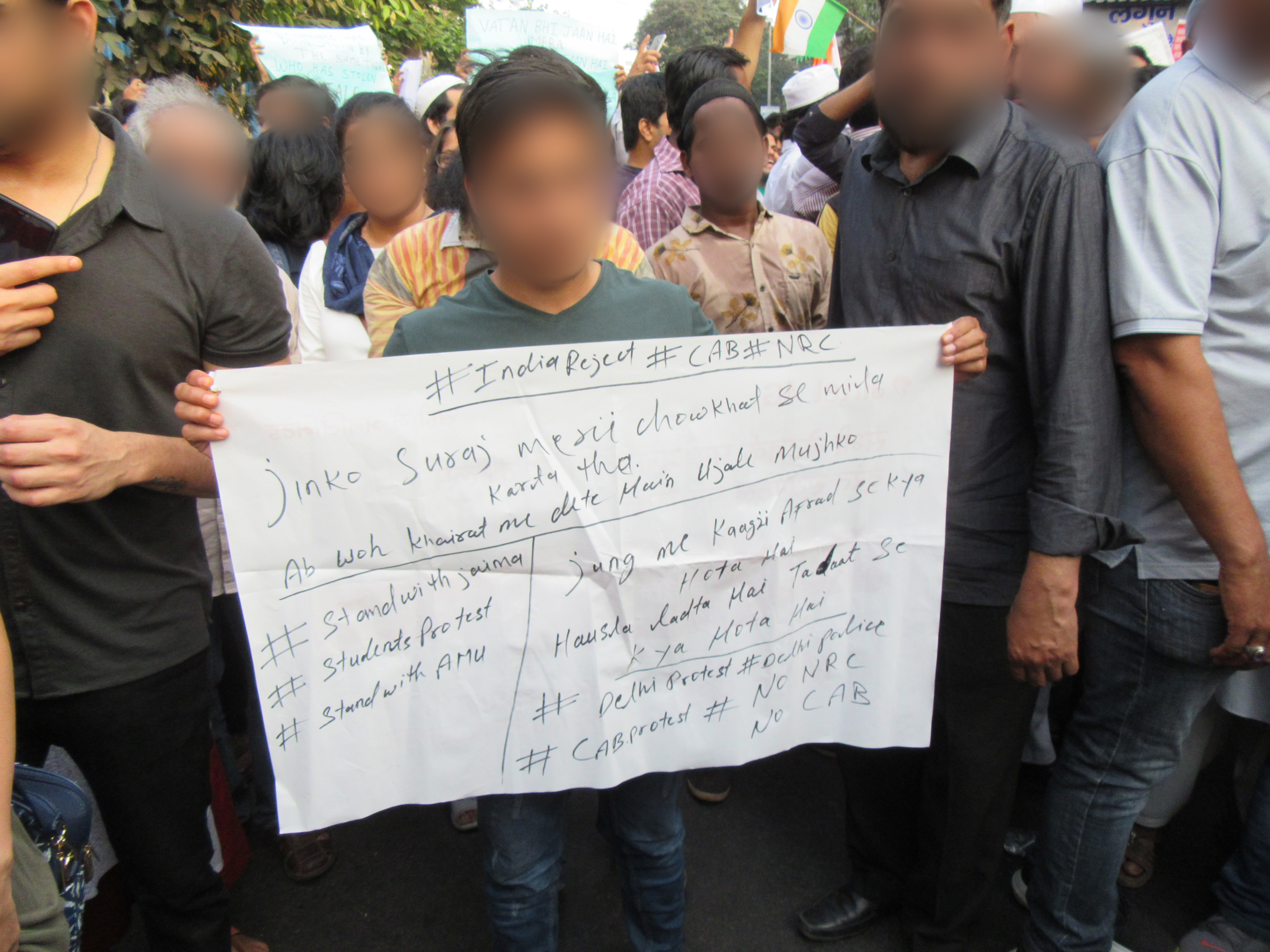 A man, standing with other protesters, holds a large handmade sign, ink on poster paper.