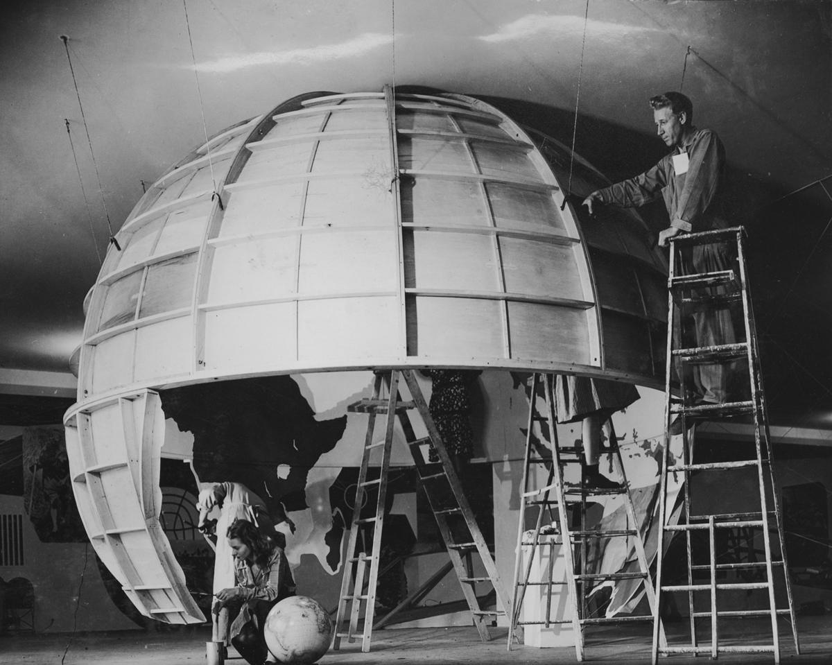 Photograph looking inside a plywood model of a globe suspended from the ceiling, with five people working and painting a world map inside.