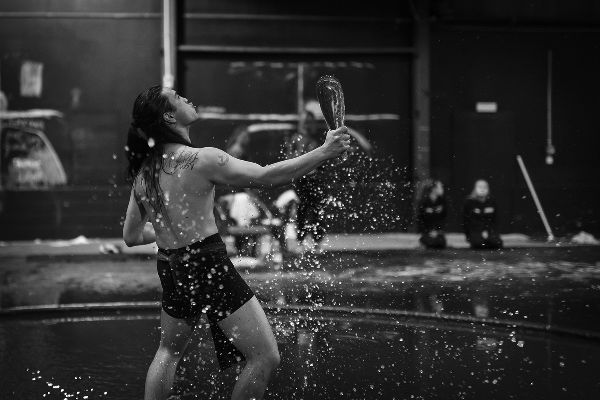 Droplets sparkle about man in black shorts with long hair who stands bent-kneed in water pool, leaning back, gaze up, arm out clenching rounded club.