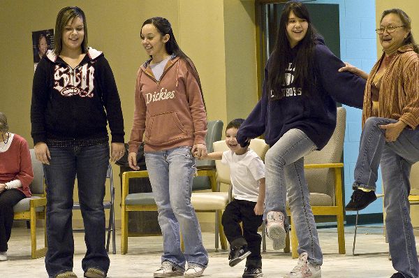 Three young women in jeans and sweatshirts stand in a row. They hold onto an older woman and a young boy, each with a leg lifted. All are laughing.