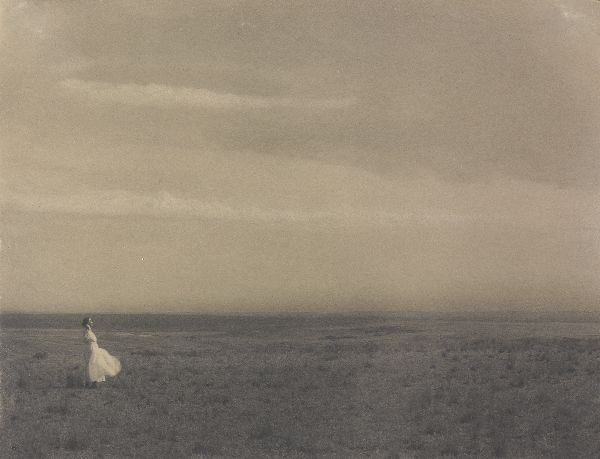 A woman in a white dress stands in a prairie landscape, facing into the frame from the left, the top of her head level with the flat horizon.