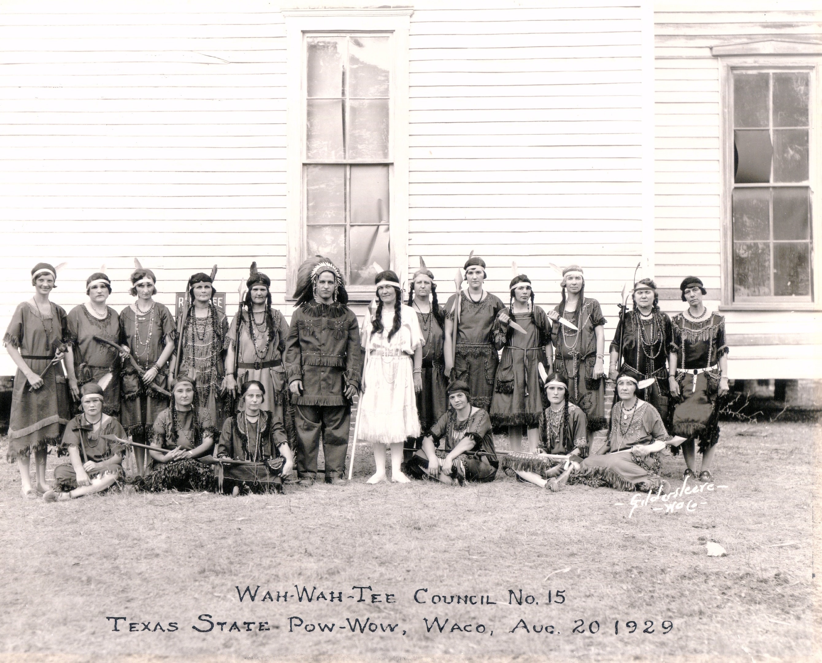 A group of white women and one white man are posed wearing Native American costumes. Many are brandishing Native American weapons or hunting tools.
