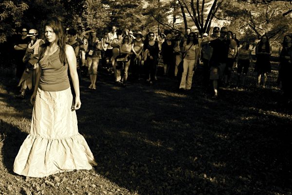 A woman in a long dress, arms down, is walking forward with a blank expression. Behind her, keeping some distance, a group of audience members watch.