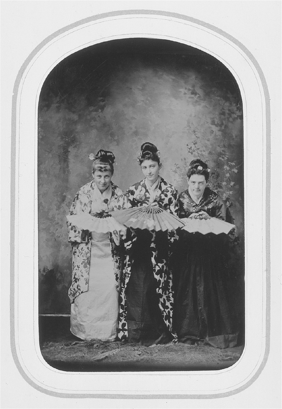 A photograph of three women in traditional attire standing together besides each other with their postures bent a little. Their hair is tied up into a high bun and each of them are holding a shan in front of them.