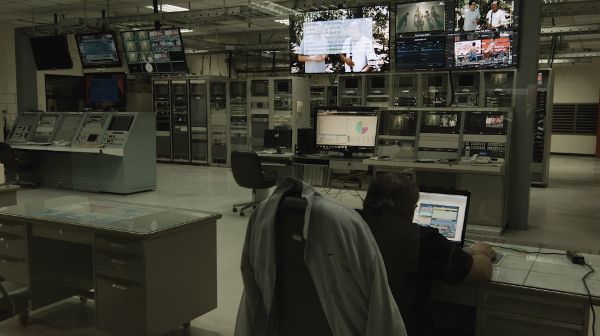 A room full of technical control panels, computing units, and monitors. Some screens are on. A man is sitting by one screen.