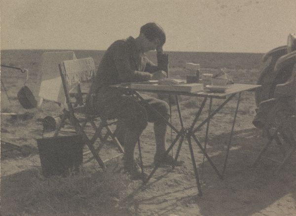 A woman in breeches, hair cut in a bob, sits on a folding chair, at a folding table. Leaning her head on her arm, she is writing.
