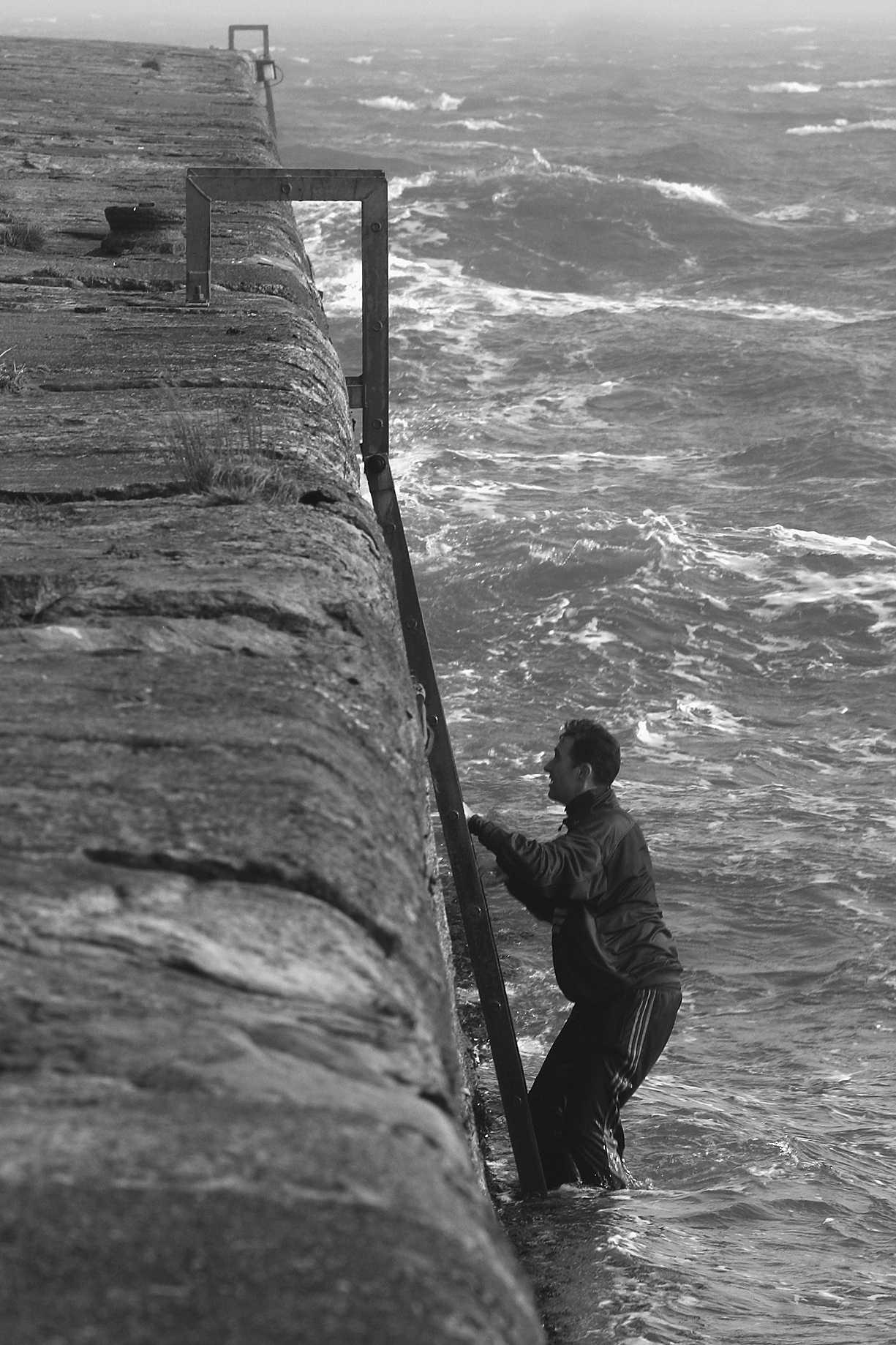 Marcus Coates climbs a metal ladder attached to a stone seawall, with rough ocean waves in the background.