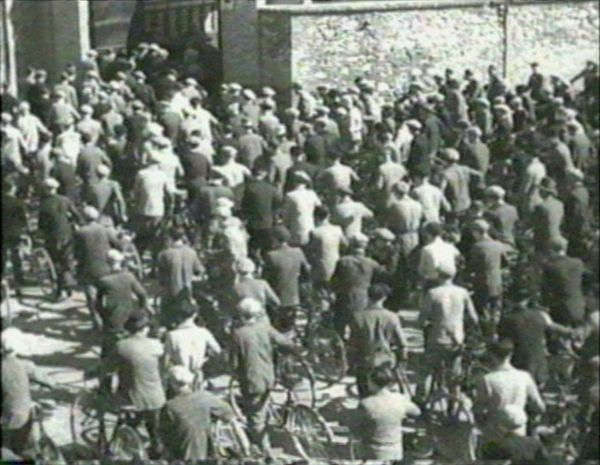 Hundreds of men, specifically factory workers, arrive to work on bicycle and enter a building.