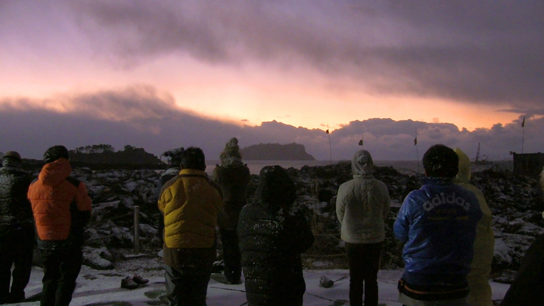 Shot from behind, nine onlookers in winter wear look out over an expanse of water toward an island in the distance, shrouded by clouds on the horizon.