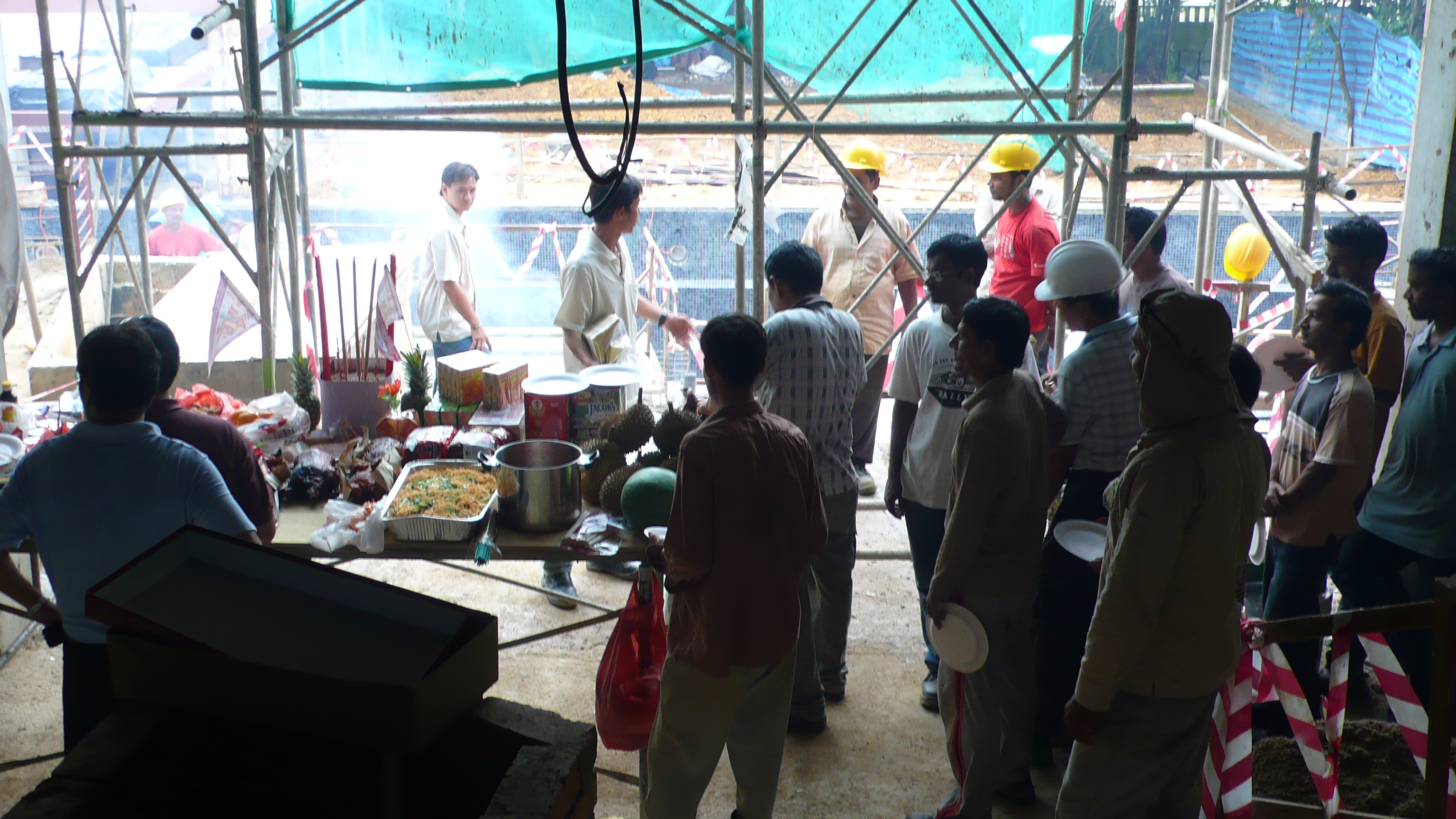 A group of men, of different Asian and South Asian ethnicities, stand near a table in a half-built house. The table contains sacrificial items.
