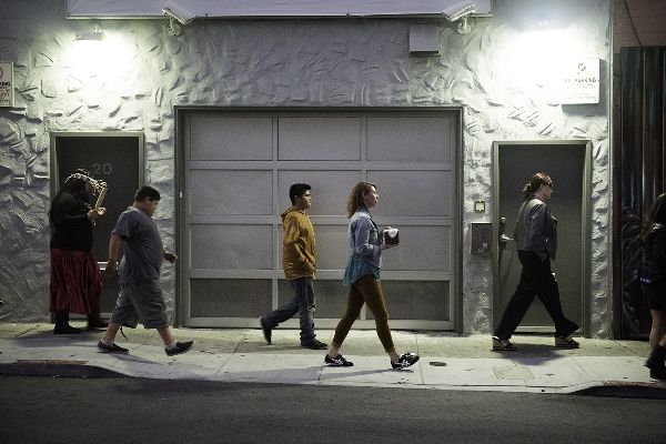 On a lit up street, in profile before a garage door, five people walk. One holds a small box. The last one, in a long skirt, raises a clapper stick.