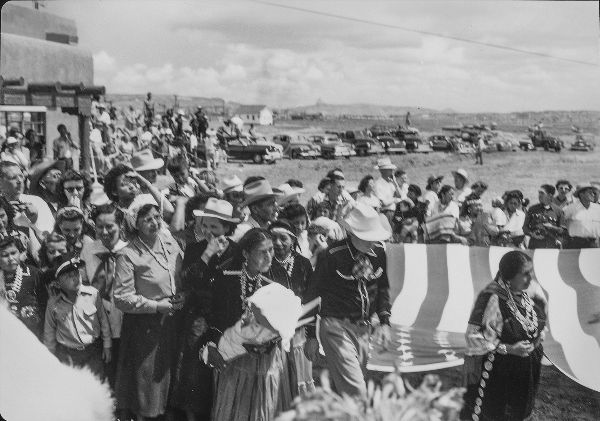 Women carry a large United States flag horizontally, in front of a crowd of people outside a building.