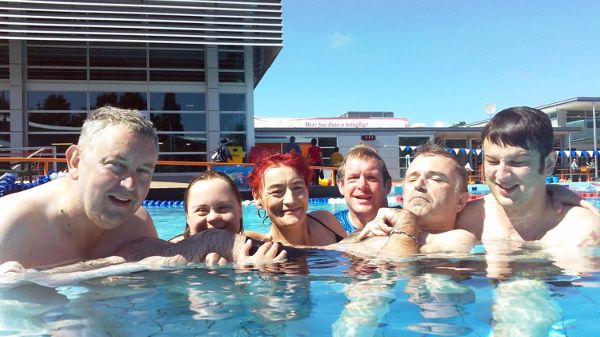 Six people in a pool looking toward the camera. One of the six is being supported by the others.