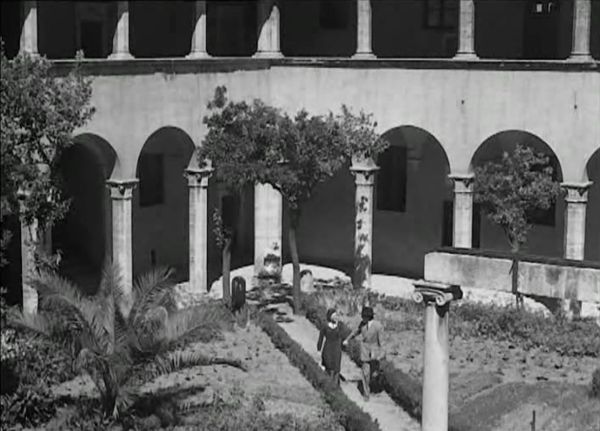 A man and woman walk hand in hand in the middle of a cloistered courtyard of an orphanage, surrounded by trees, plants, and an ancient column.