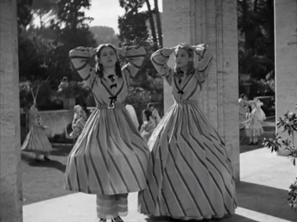 Two young women in long, striped dresses stand outside with their arms crossed behind their heads as similarly dressed women parade behind them.