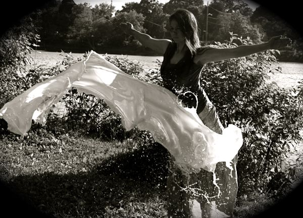 A woman is standing with arms out, leaning forward, near a river. A sheet of white milk, hurled at her, hangs in the air and is about to smack her.