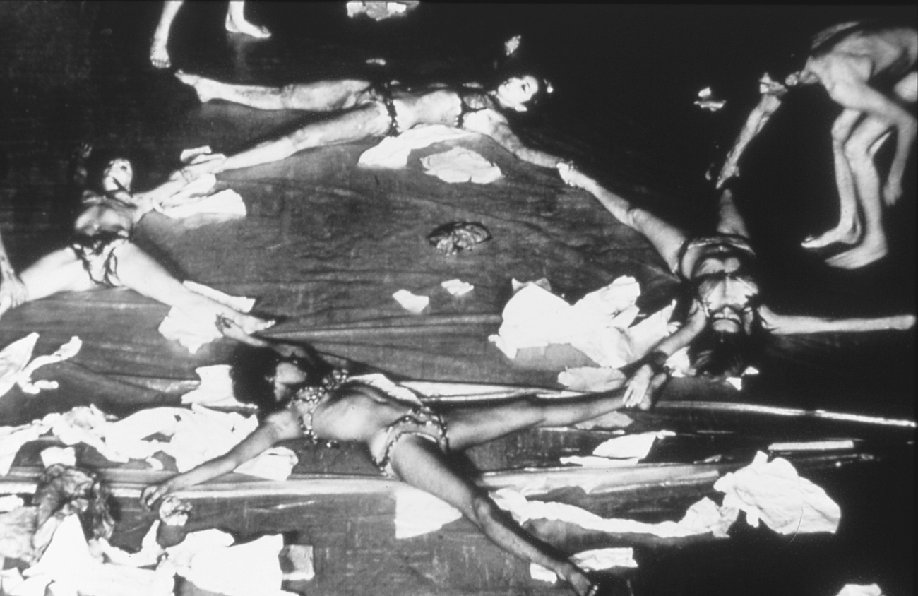 A performance featuring several individuals, Carolee Schneemann, Annina Nosei, Stanley Gochenouer, Sandra Chew, and Dorothea Rockburne, lying on a stage in dynamic, spread-out poses, surrounded by scattered paper and fabric.