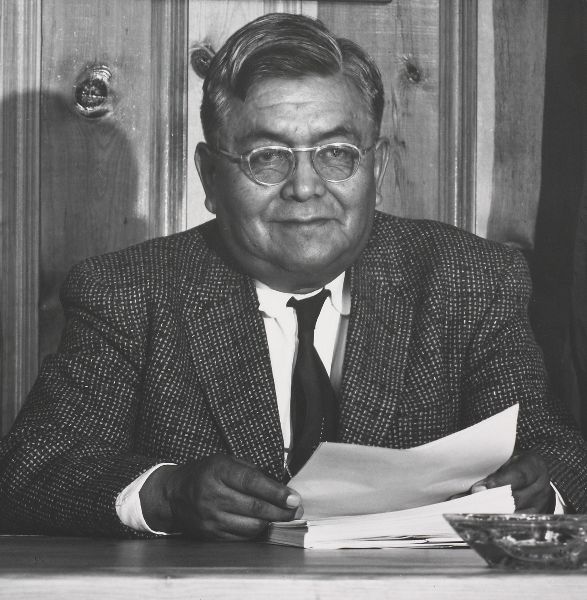A man with glasses at a desk in front of wood paneling, holding a sheet of paper from a stack in front of him.