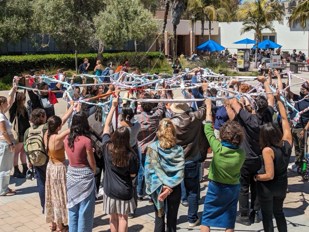 Students, faculty, and community members stand in a circle, each holding part of a long chain made out of fabric. Other students can be seen mingling in the background.