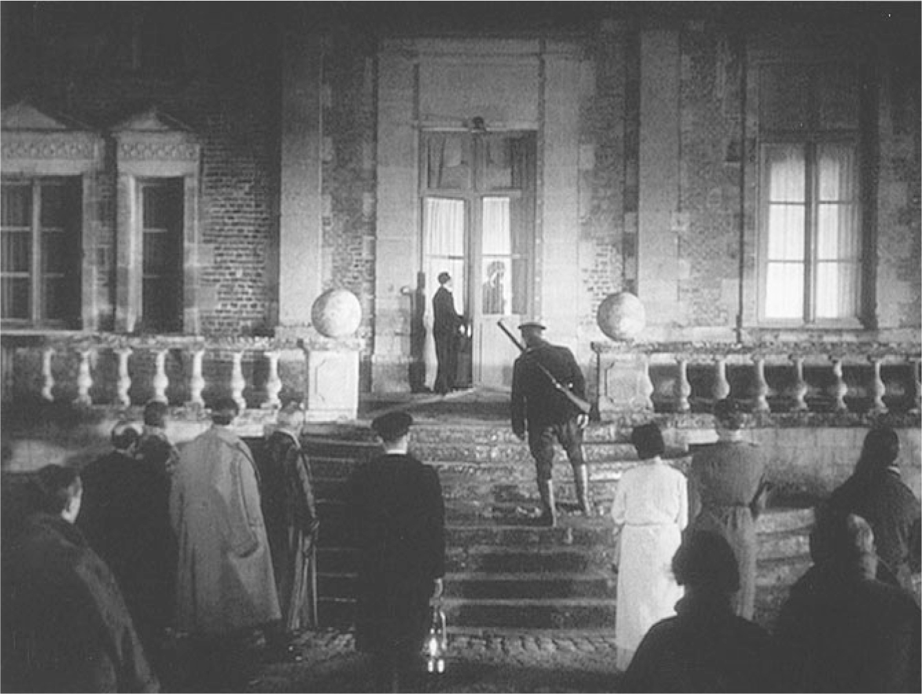The exhibit depicts a scene from “La Règle du jeu” where a group of men and women stand outside a house, with a man in the forefront holding a gun over his shoulder. At the entrance of the house, another man is seen looking just past the slightly opened door.