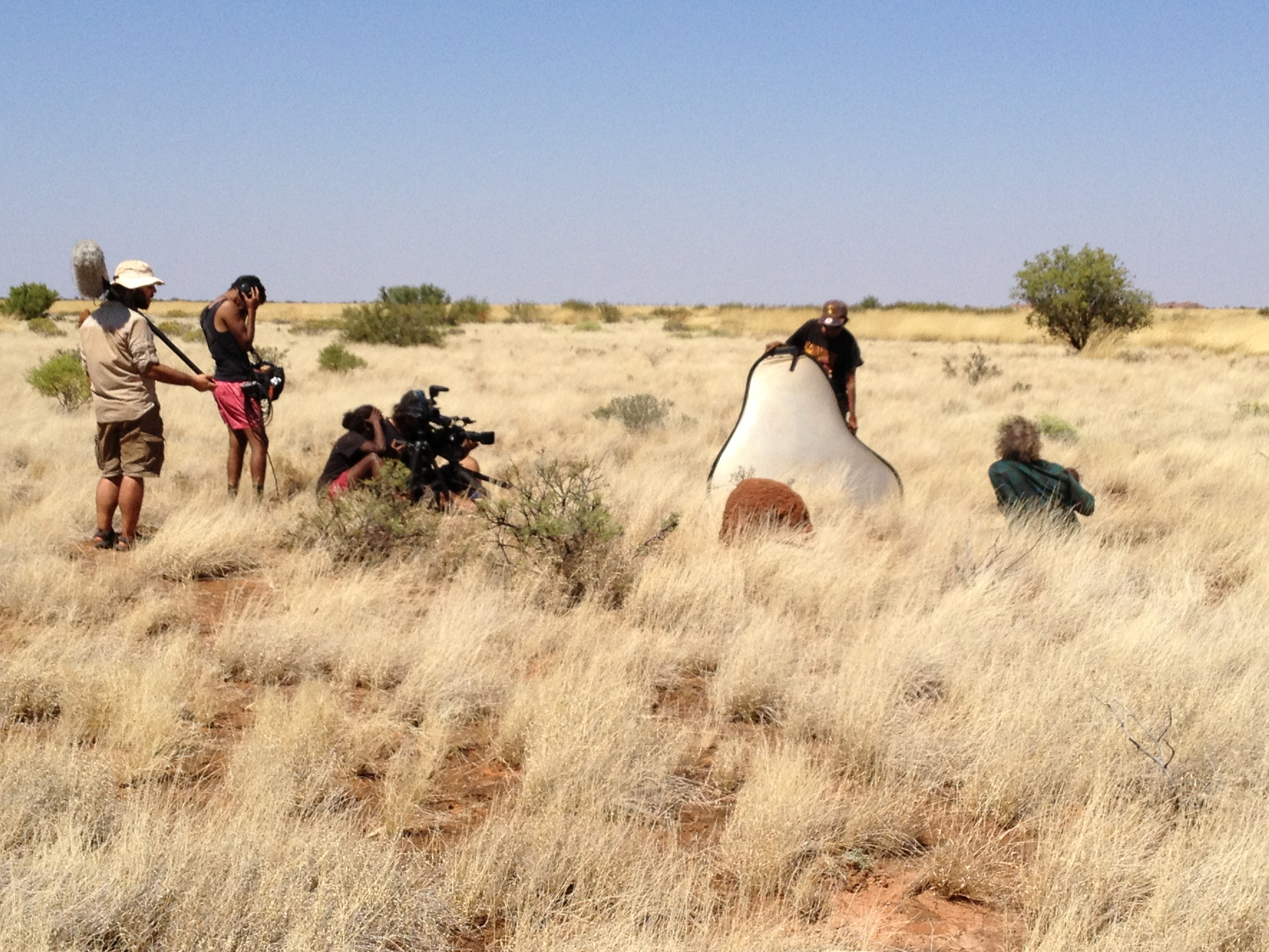 On a sunny day, Mark sits in spinifex tall grasses on the right as a camera crew gathers around him with video and audio equipment.