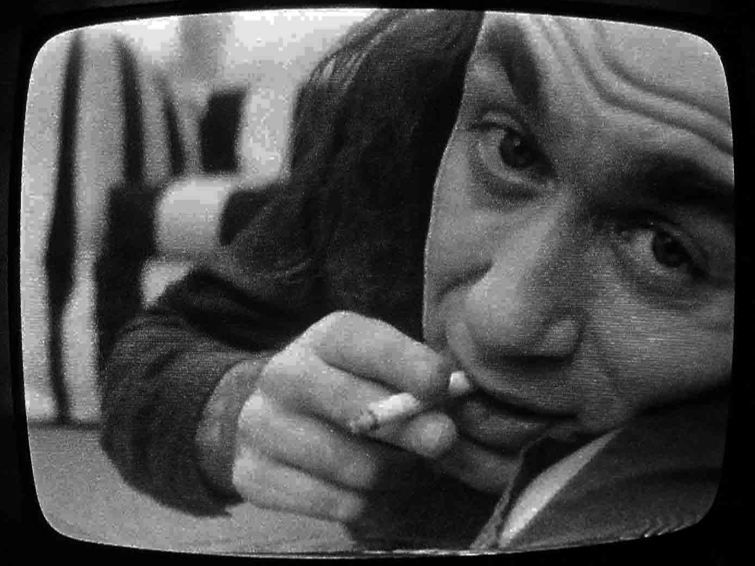 A similar still, but this time he’s holding a cigarette and the image corners are curved. A couch with vertical stripes is still visible.