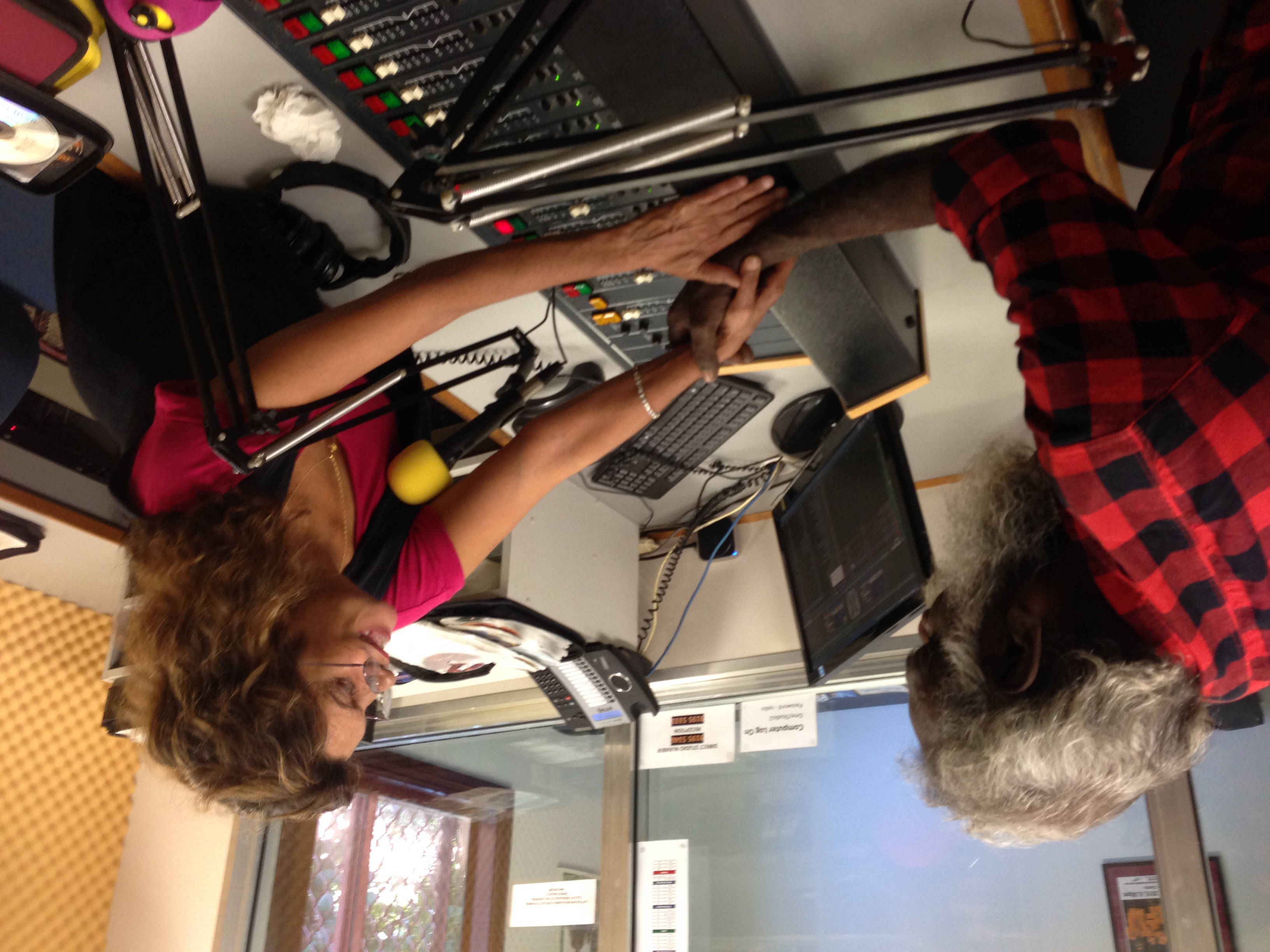 Mark Moora and Sandy Dann shake hands while smiling toward one another. Their hands reach across a radio desk with controls and microphones.