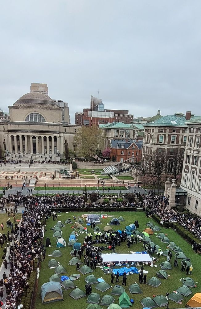 An overhead view of the Pro-Palestinian encampment on the grassy field on Columbia University campus. A group of protestors kneel in the center of the image, surrounded by NYPD officers who are standing in a circle around them. Beyond the boundaries of the tents other protests and onlookers stand clustered around the boundaries of the lawn observing the arrest.