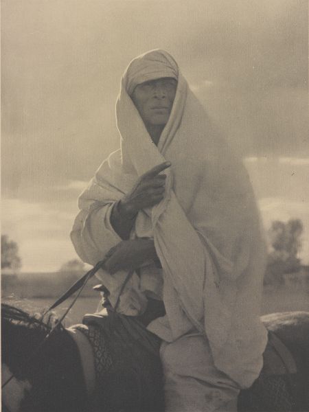 Close-up from below of a man swathed in white fabric, riding a  horse, gazing and pointing into the distance.