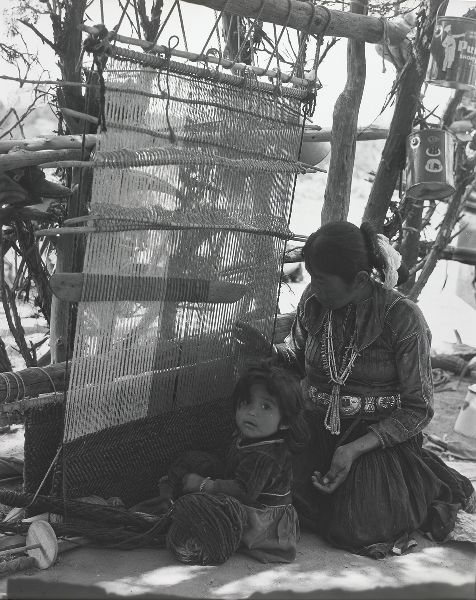 A woman and young child sit in front of a loom; the woman puts her fingers through the warp and the child looks at the camera.
