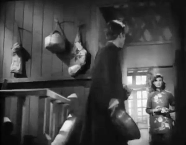 Hallway scene from behind the man’s shoulder, showing him looking toward the smoking woman who is from this vantage further down the hall.