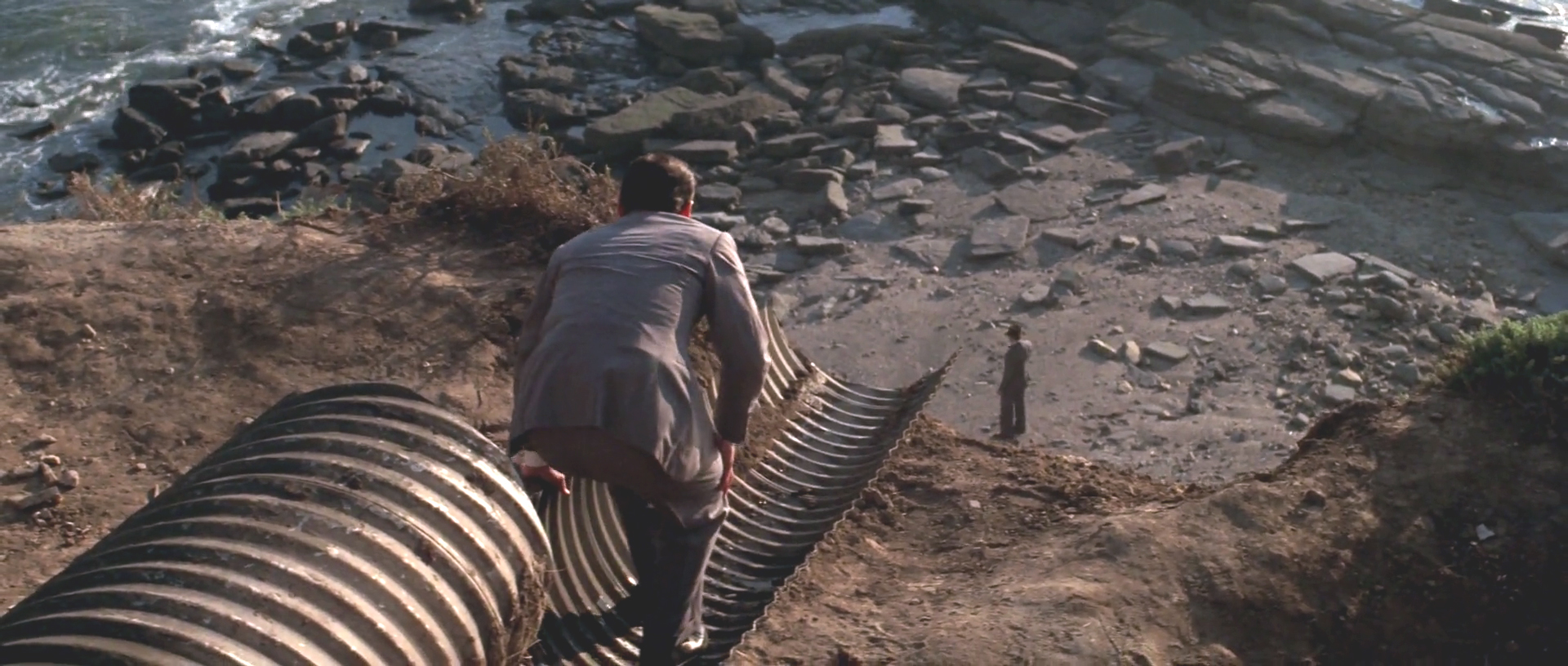 Jake stands inside a half-pipe drain looking down on Mulwray who is looking out into the ocean.