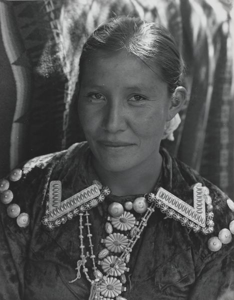 A head shot of a woman wearing a decorated blouse and silver jewelry, smiling as she makes eye contact with the camera.