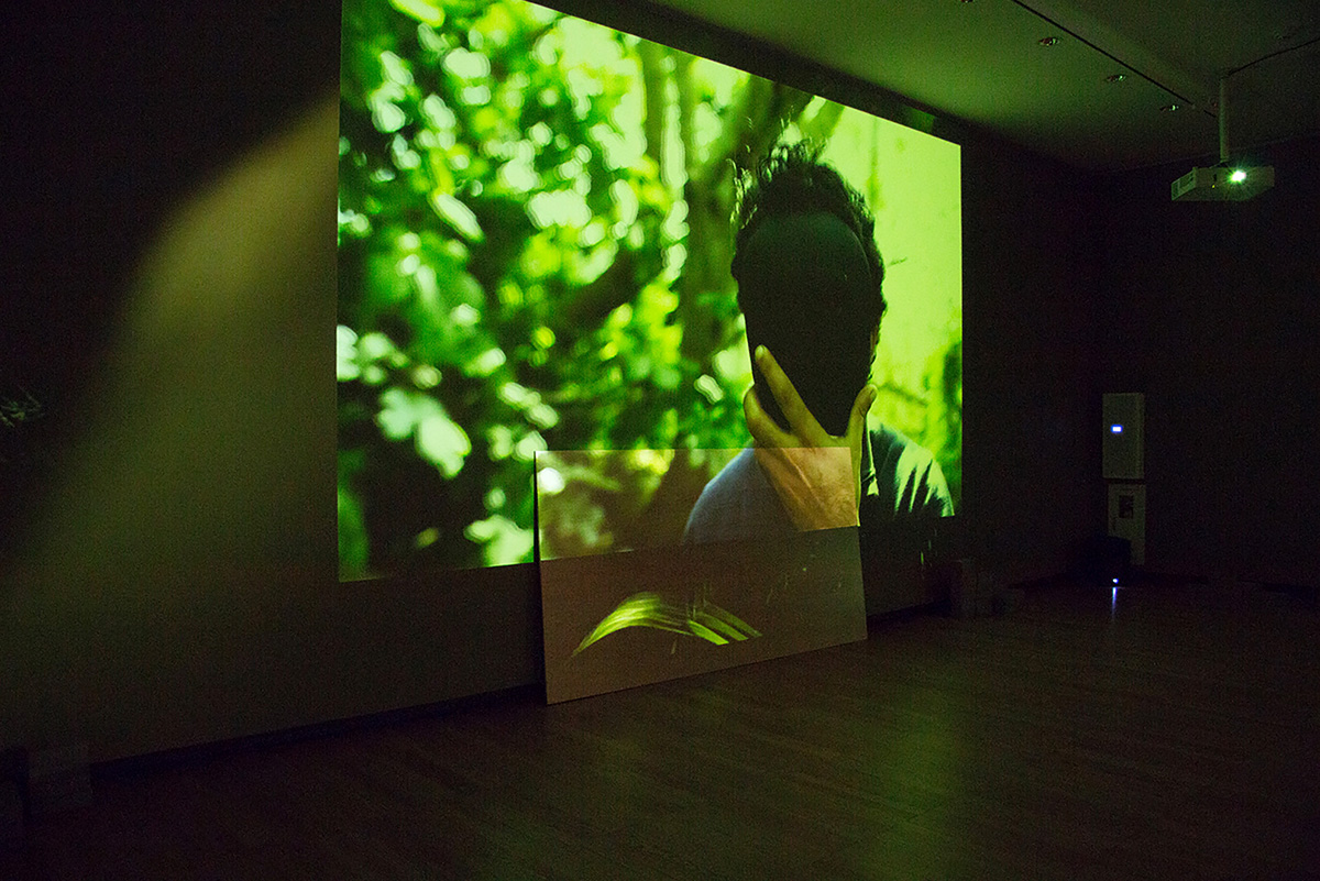 Image of video projected on a wall showing a figure standing in front of nondescript green flora, covering their face with a black, featureless mask.