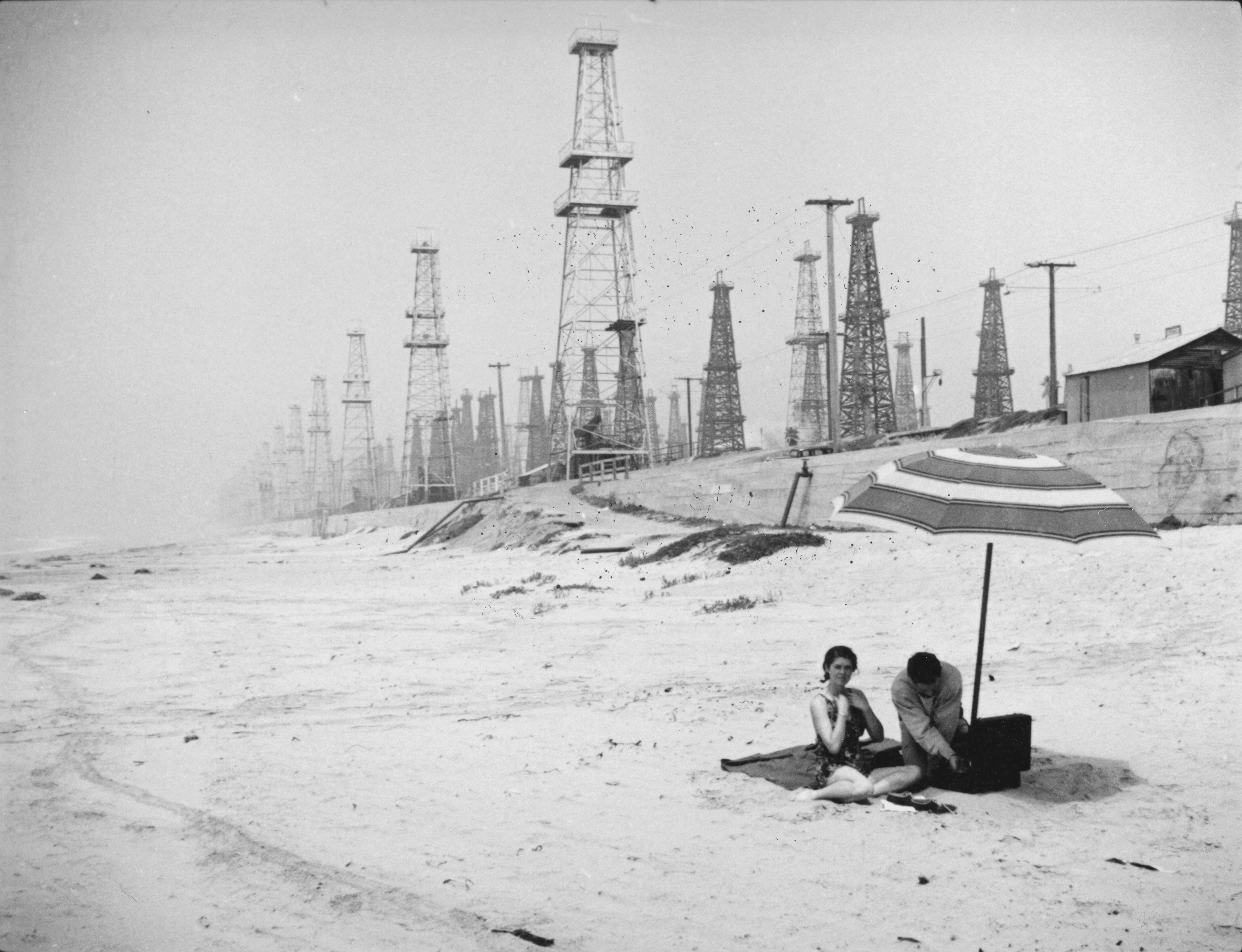 A man and a woman lounge on the beach under an umbrella. Hundreds of oil derricks are visible behind them.